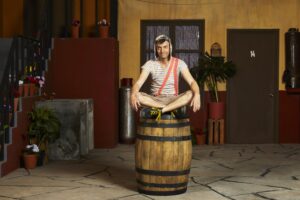 A man dressed as boy in a striped shirt, shorts and orange suspenders sits on a barrel in a courtyard.