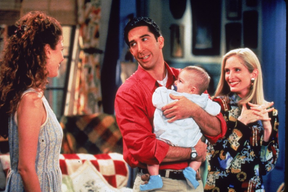 Still from *Friends* showing David Schwimmer holding a baby while Jessica Hecht and Jane Sibbett look on.
