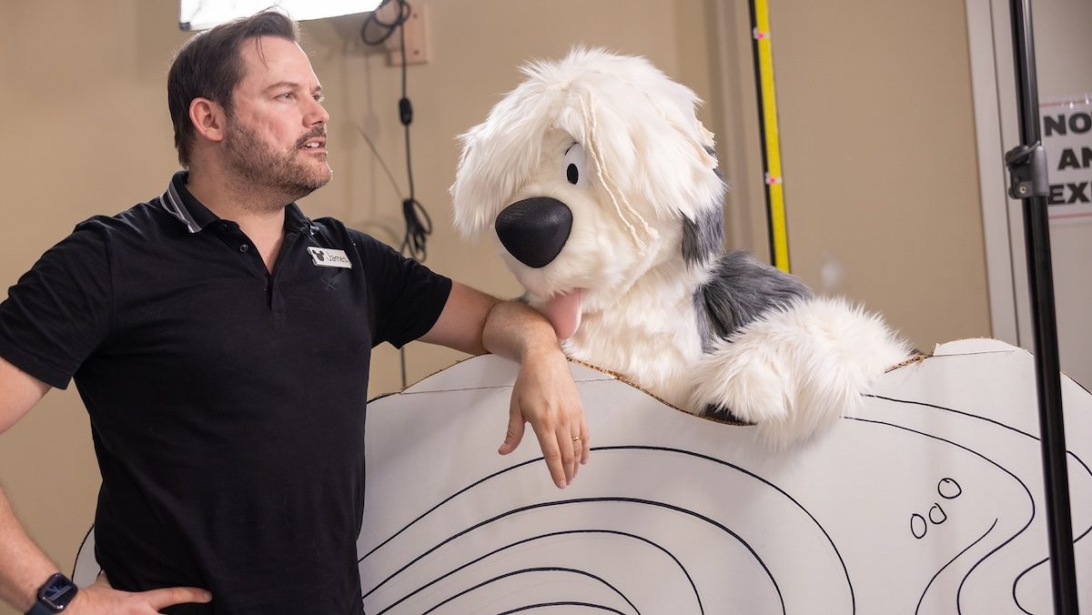 A man in a black golf shirt leaning near a puppet of the dog Max from The Little Mermaid