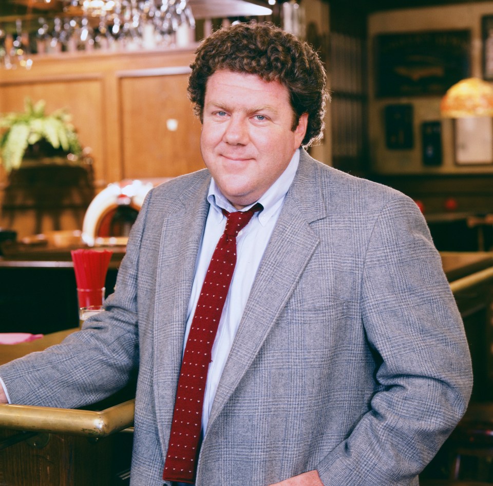 George Wendt as Norm Peterson in a gray jacket and red tie.