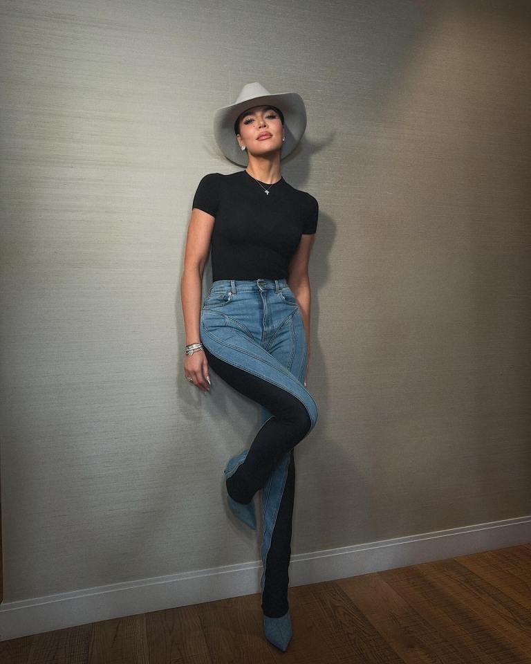 Khloe Kardashian wearing a black top, light wash jeans with black paneling, and a light-colored cowboy hat.