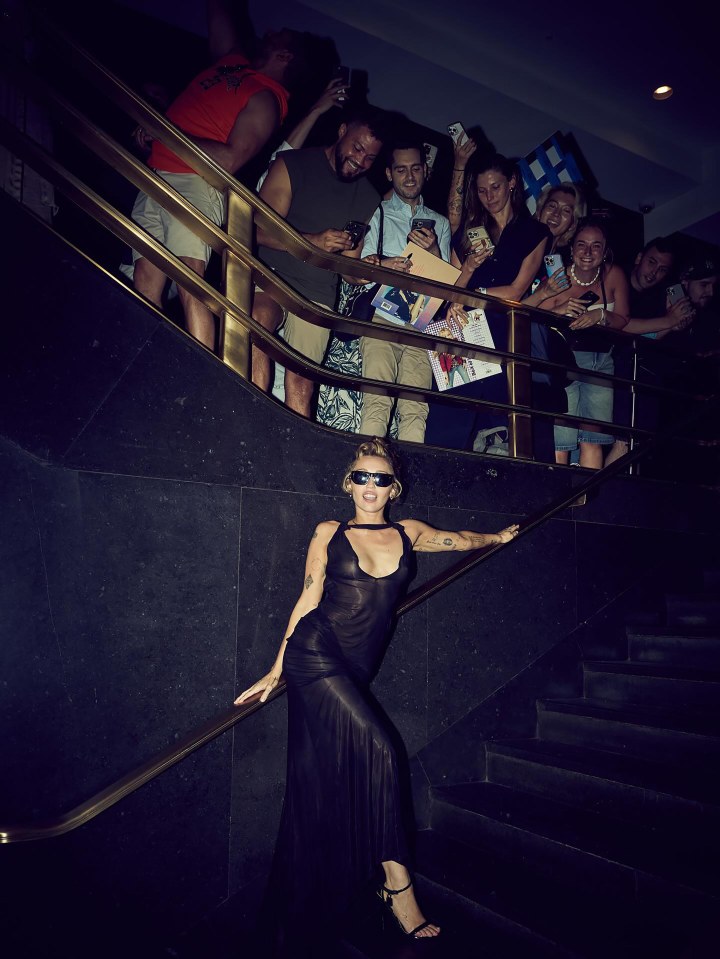 Miley Cyrus in a sheer black dress on a staircase, surrounded by fans.
