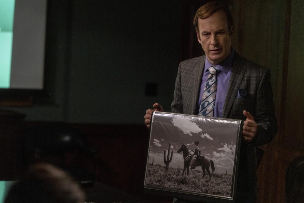 Bob Odenkirk, in a suit, holds a picture of a person on horseback in the desert