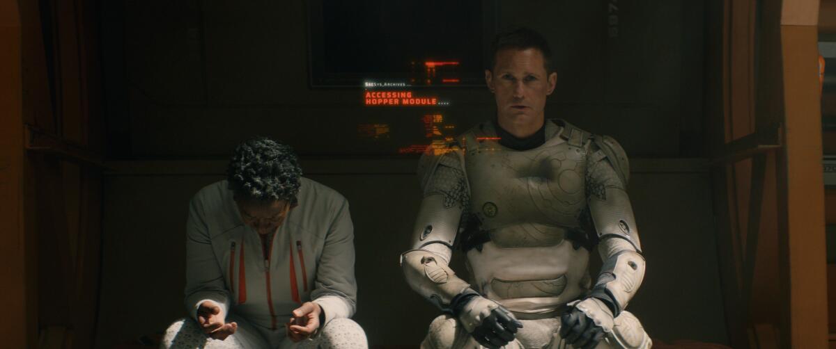 A woman (Mensah) sitting with her head down next to a person in white armor (Murderbot) who is looking straight ahead.