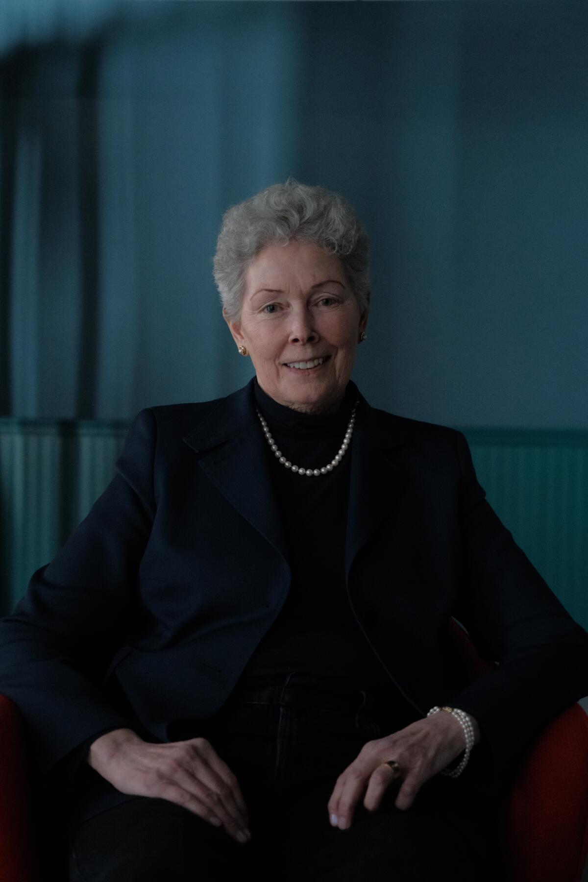 A woman with short grey hair in a dark turtleneck and blazer sitting.