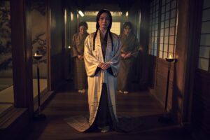 A woman in a white kimono flanked by two women in green kimonos.