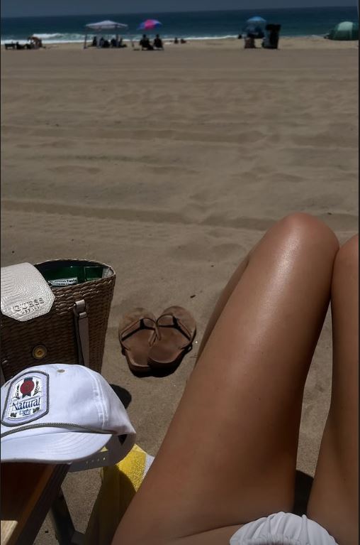 Person's legs on a beach with sandals, a hat, and a bag.
