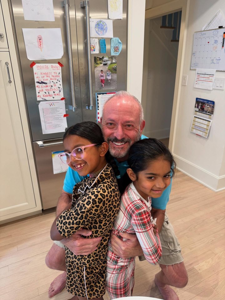 A man hugging two girls.  House rules are visible on a refrigerator in the background.