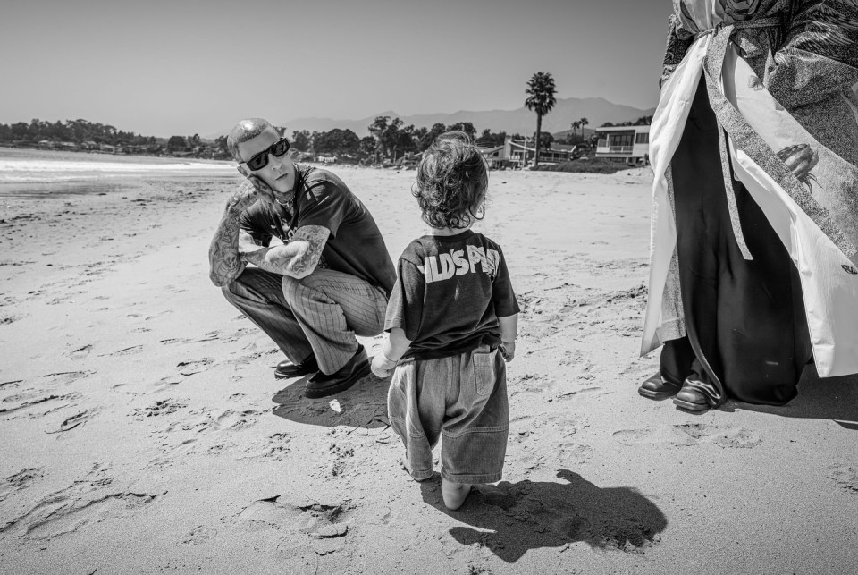 Black and white photo of a man with tattoos crouching on a beach, looking at a toddler.  A person in a robe is visible in the background.