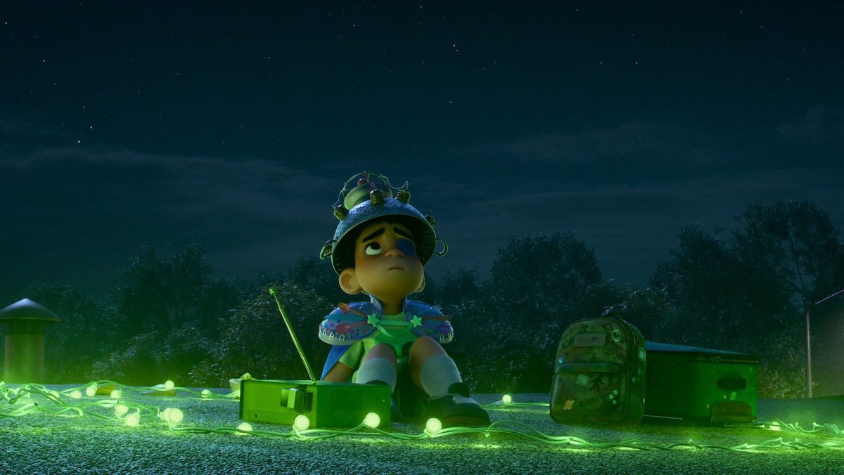 A kid in a weird costume and sieve on his head sits among green lights looking up at the night sky in Elio