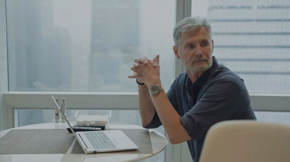 A man sits at a table with a laptop, looking away from the camera.