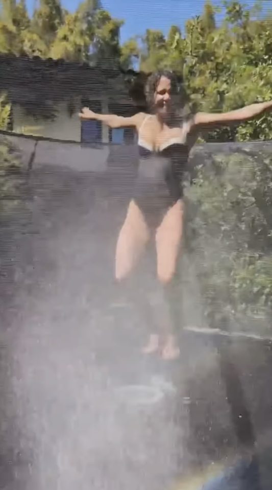 Woman in swimsuit bouncing on trampoline while being sprayed with water.