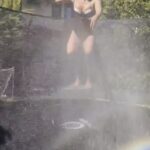 Woman in a swimsuit bouncing on a trampoline while being sprayed with water.