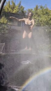Woman in a swimsuit bouncing on a trampoline while being sprayed with water.