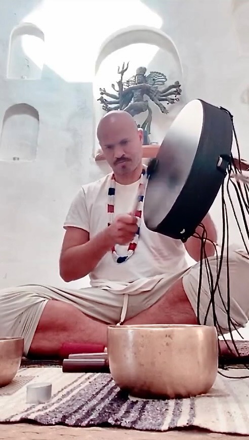 Man playing a large drum while sitting cross-legged, surrounded by singing bowls.
