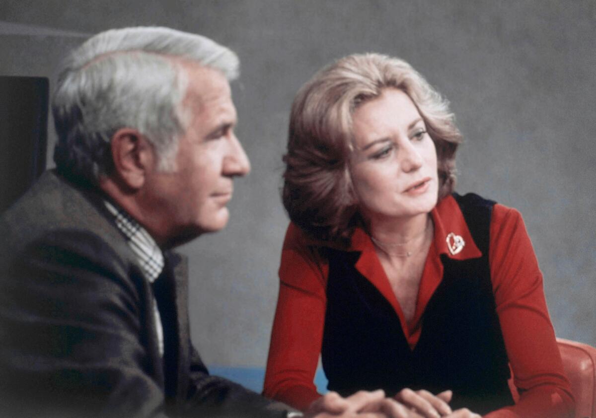 A profile view of a man with white hair sitting next to a woman in a red top and black vest.