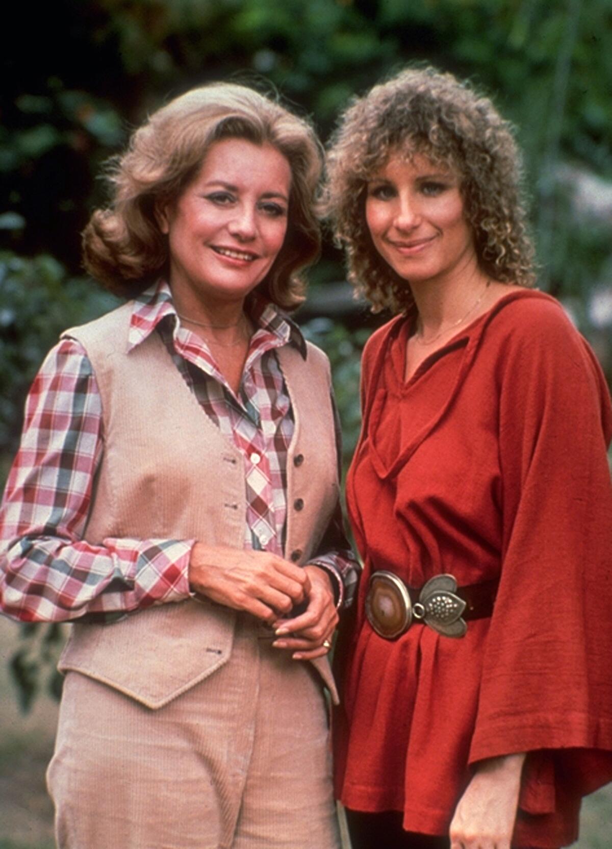 A woman in a tan vest, slacks and plaid shirt stands next to a woman in a red dress.