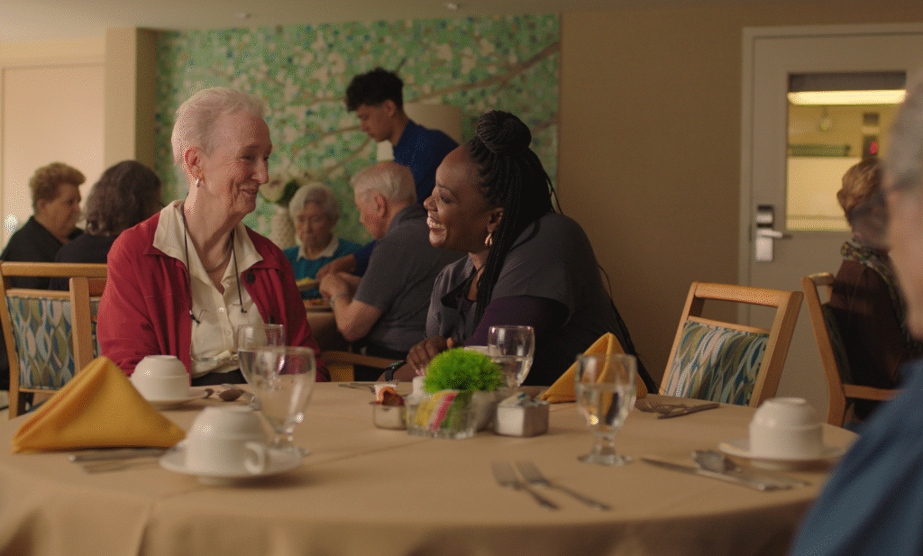 A woman sits at a dining table with another welcoming person.