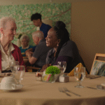 A woman sits at a dining table with another welcoming person.
