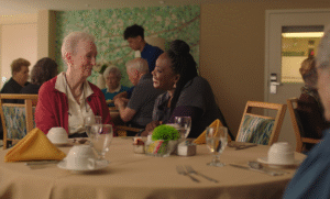 A woman sits at a dining table with another welcoming person.