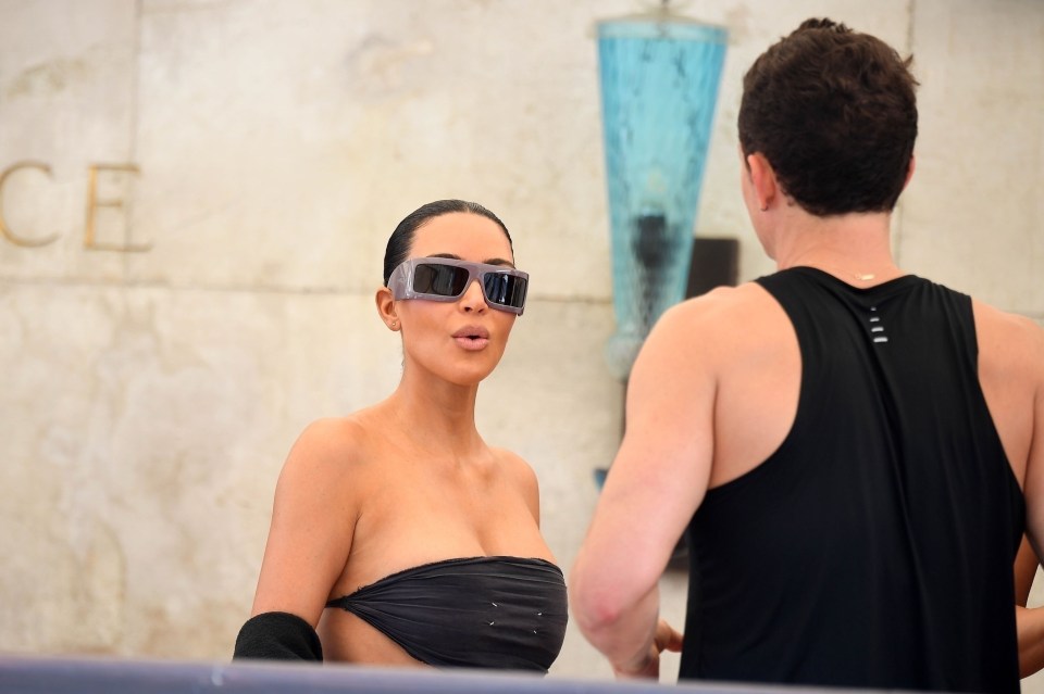 Kim Kardashian wearing sunglasses, talking to a person wearing a black tank top.