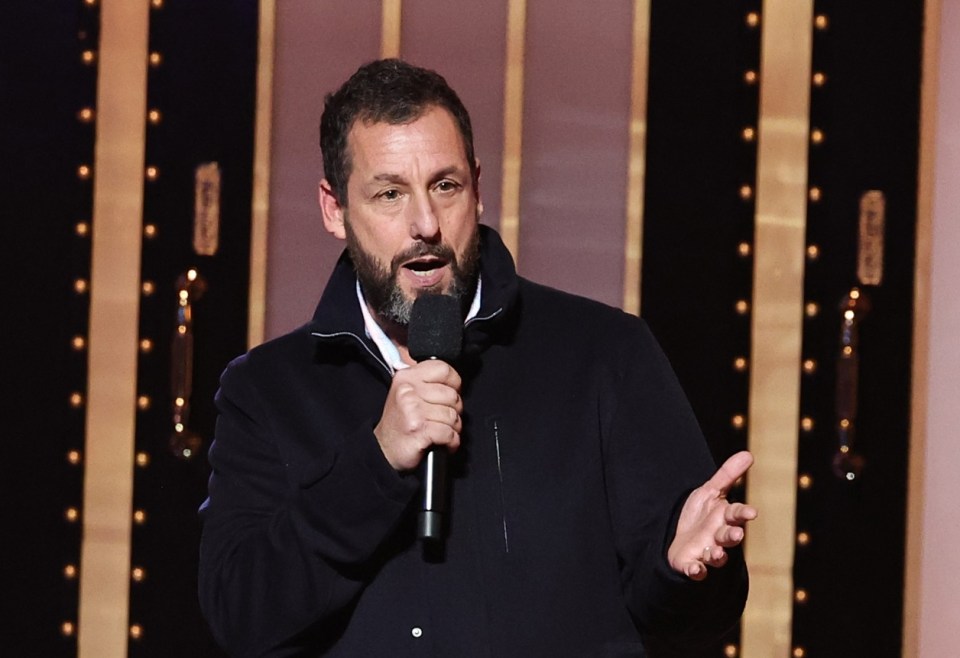 Adam Sandler speaking into a microphone on stage.