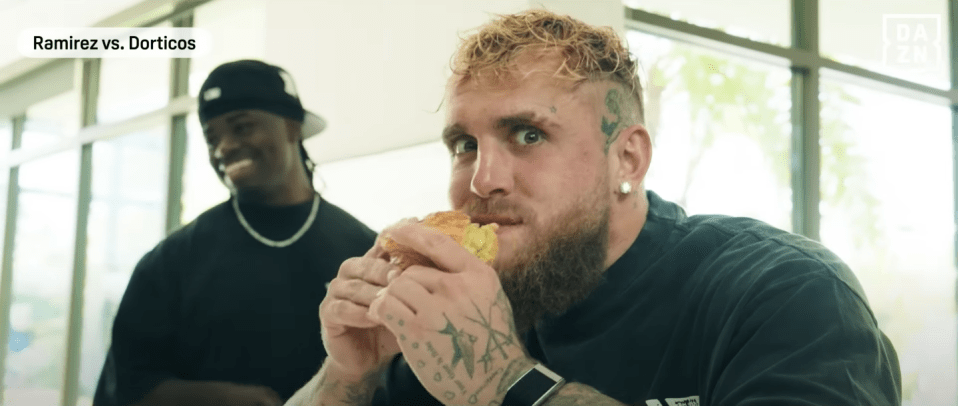 Man eating a sandwich with another man in the background.