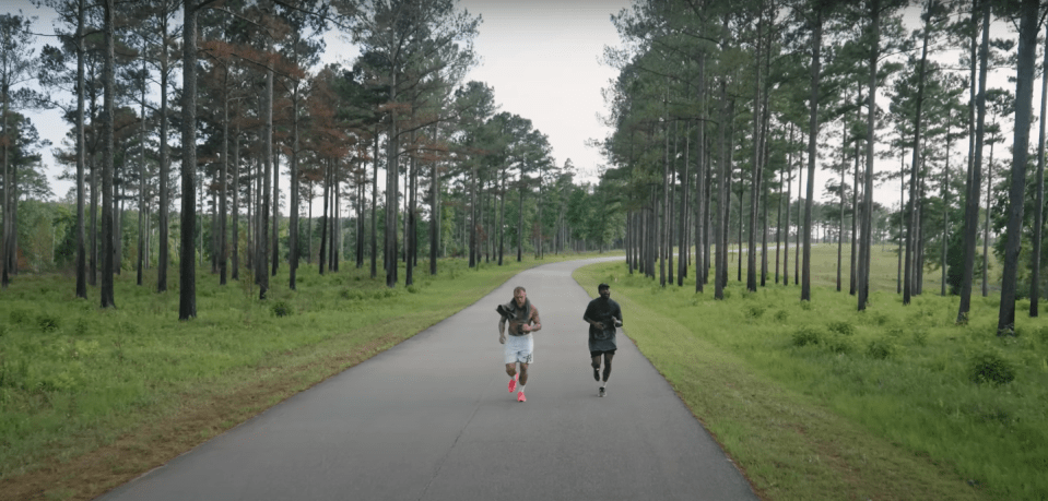 Two men jogging on a paved road lined with tall pine trees.