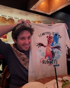 Person wearing a tiara and sash holding up a shirt featuring a cartoon rooster; the shirt says, "I can feel it in my Nuggets."
