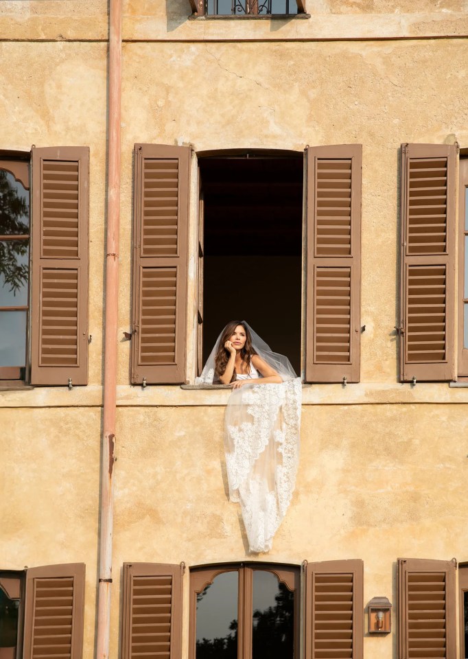 Bride in wedding dress and veil looking out window of old building.