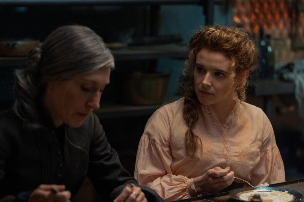 A woman with greying hair sits eating next to a woman with curly red hair in a pink top.