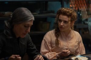 A woman with greying hair sits eating next to a woman with curly red hair in a pink top.