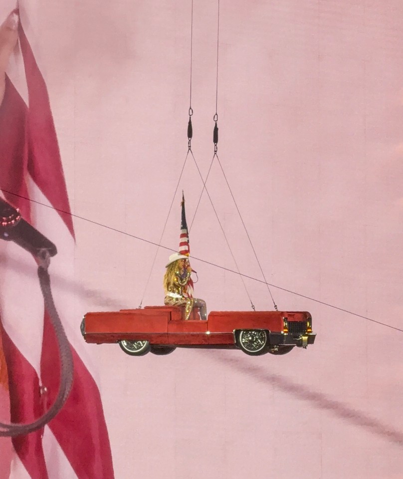 Beyoncé in a red car suspended from the ceiling, holding an American flag.