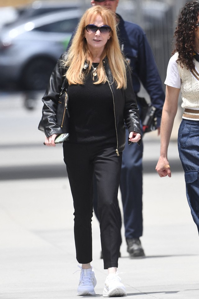 Rebecca De Mornay walking outside wearing a black leather jacket, black pants, and white sneakers.
