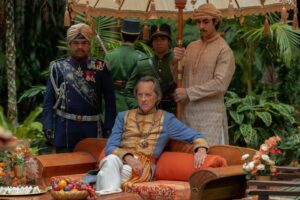 Richard E Grant as The Rajah sits on a couch surrounded by guards and servants in Nautilus