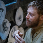 Harry (Alan Tudyk) looks sadly at sketches of Elizabeth Bowen’s Deputy Liv Baker and Corey Reynolds’ Sheriff Mike Thompson in season 4 of Resident Alien