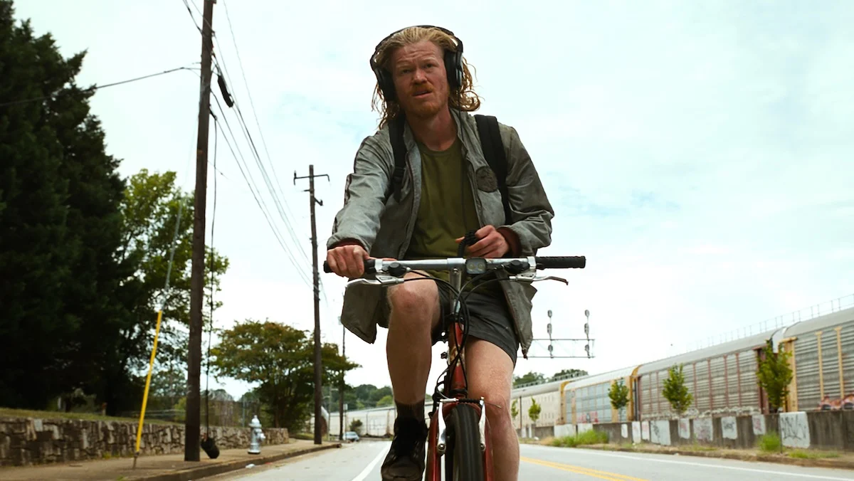 A gaunt long haired scuzzy Jesse Plemons riding a bike in Bugonia