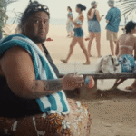 A large person with tattoos sits on a bench at the beach, holding a drink.  People are walking by in the background.