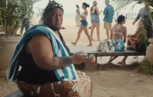 A large person with tattoos sits on a bench at the beach, holding a drink.  People are walking by in the background.