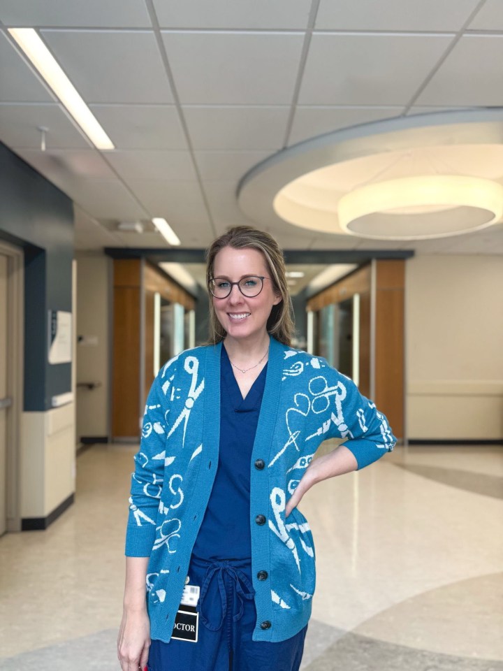 A doctor wearing scrubs and a teal cardigan with medical instrument designs stands in a hospital hallway.