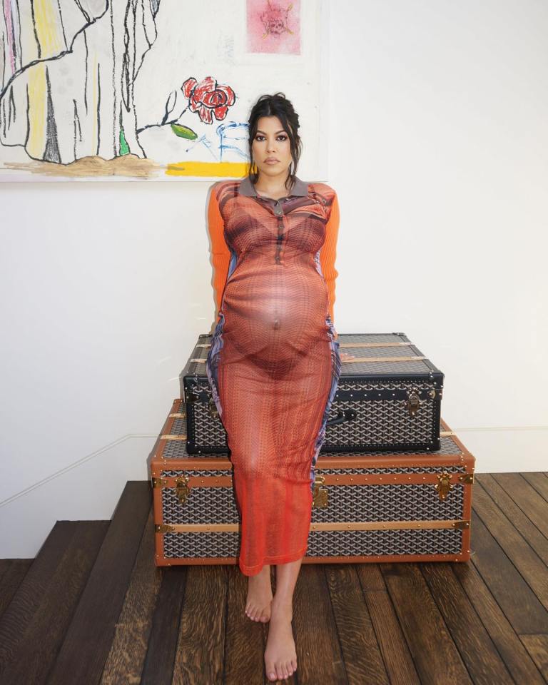 Kourtney Kardashian, pregnant, in orange dress, sitting on luggage.