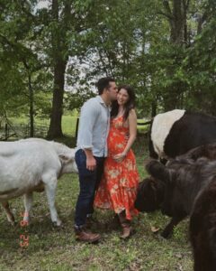Pregnant woman in orange dress being kissed by a man, with cattle nearby.
