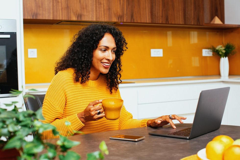Retrato de uma mulher com cabelo preto e encaracolado sorrindo enquanto usa o computador laptop na cozinha. Ela está usando um suéter amarelo-laranja que combina com a cor da parede