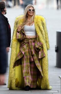 Fergie on the set of the Netflix film *Toomuch*, wearing a yellow fur coat and plaid pants.