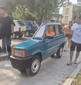 A teal, ultra-thin car, narrower than a motorcycle, being driven down a street.