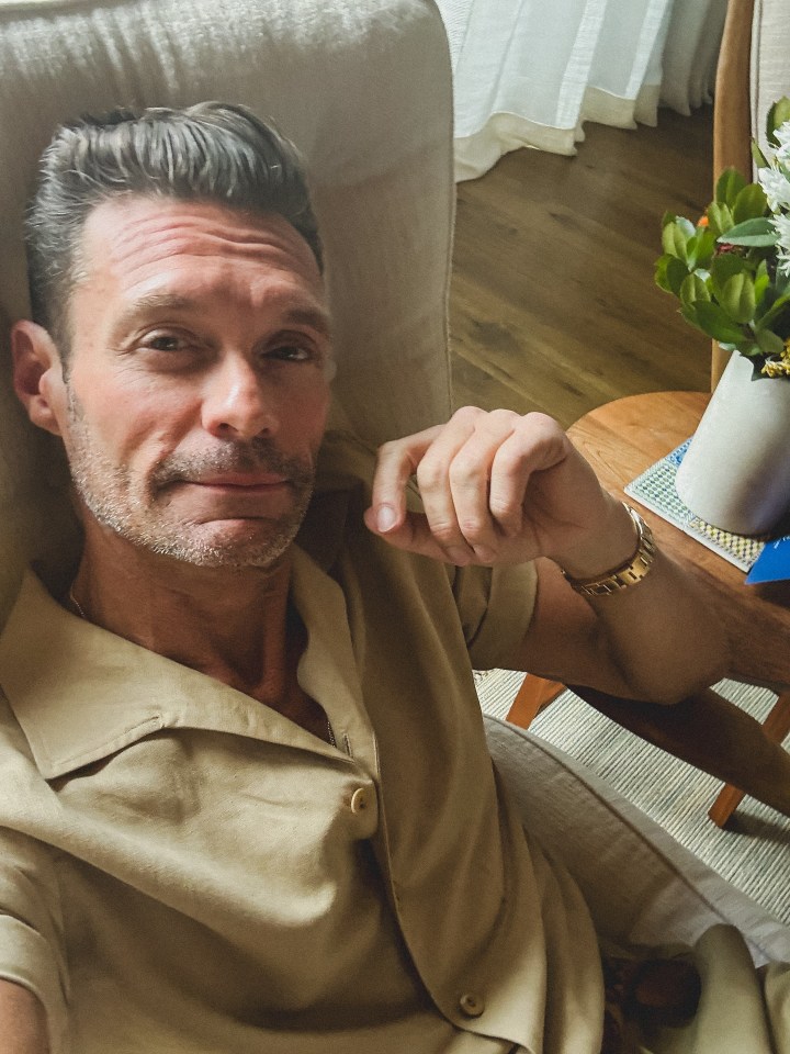 Ryan Seacrest in a beige shirt, relaxing in a chair.