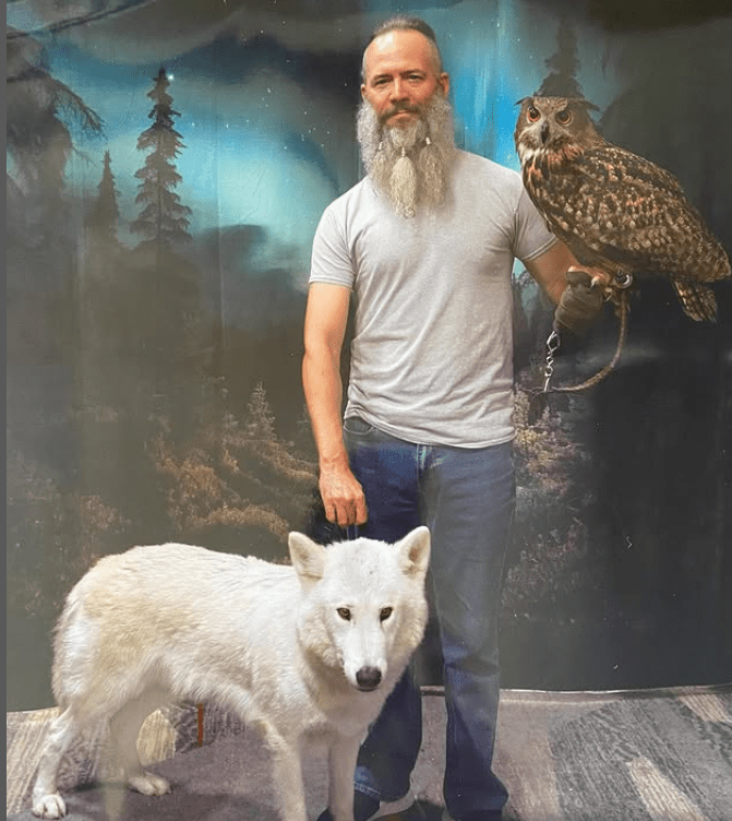 Man with long beard holding an owl and standing next to a white wolf.