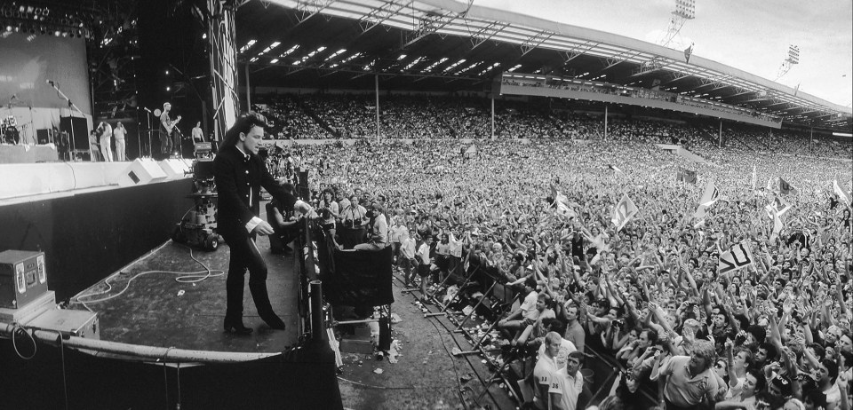 Black and white photo of a performer on stage in front of a large crowd at a stadium concert.