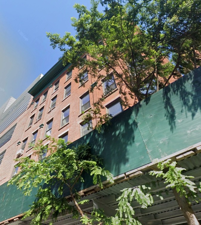 Brick building partially obscured by construction barrier and trees.