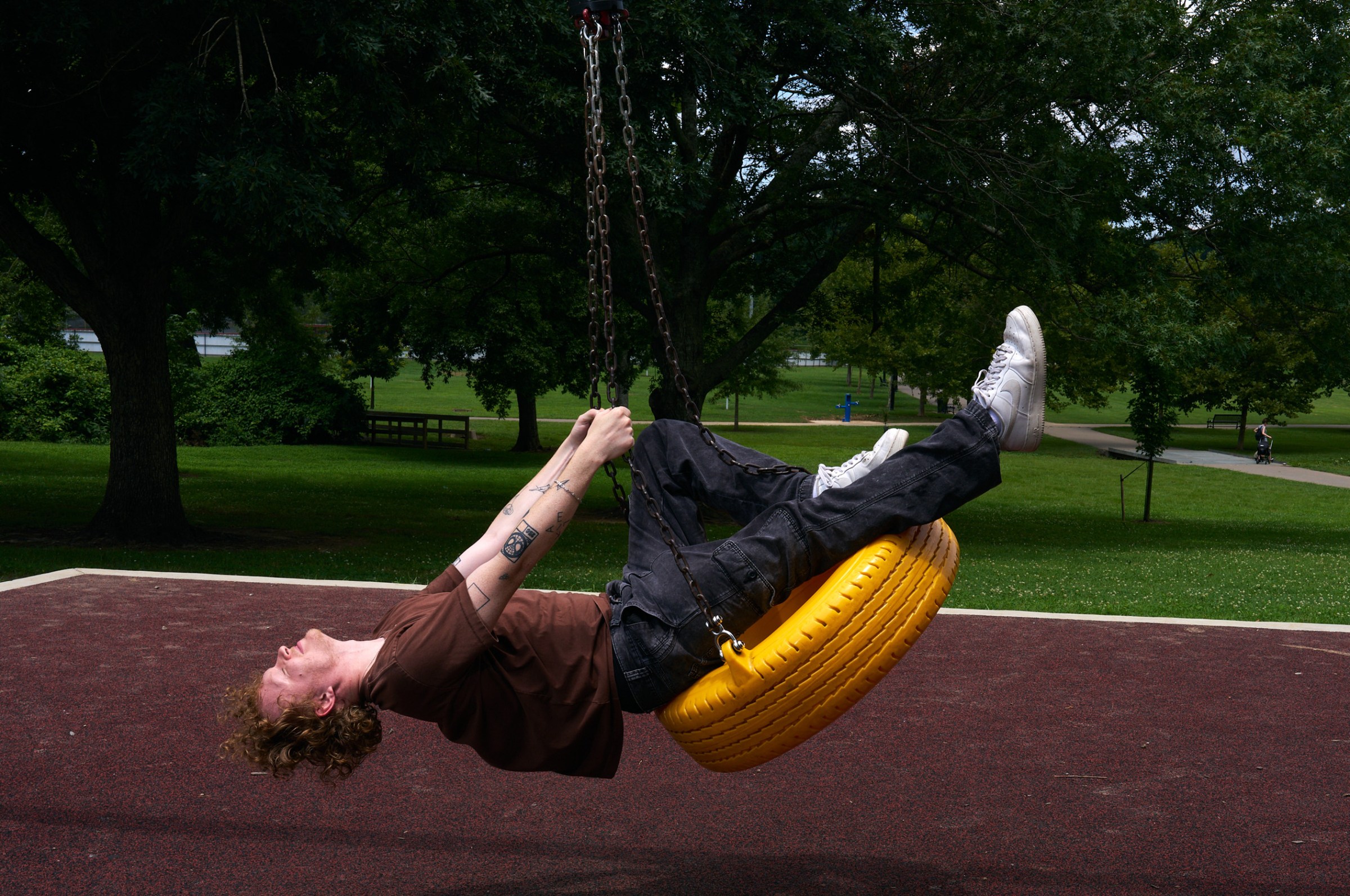 Knox leans back with eyes closed and arms extended while swinging on a bright yellow tire swing. The ground beneath him is brown and behind is an expanse of a bright green park lawn.